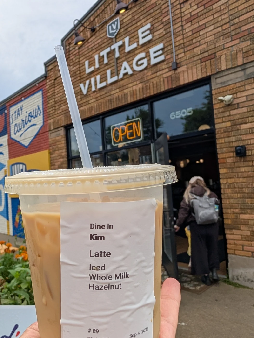 Kim’s iced latte held up outside the Little Village café, with its sign above the door.